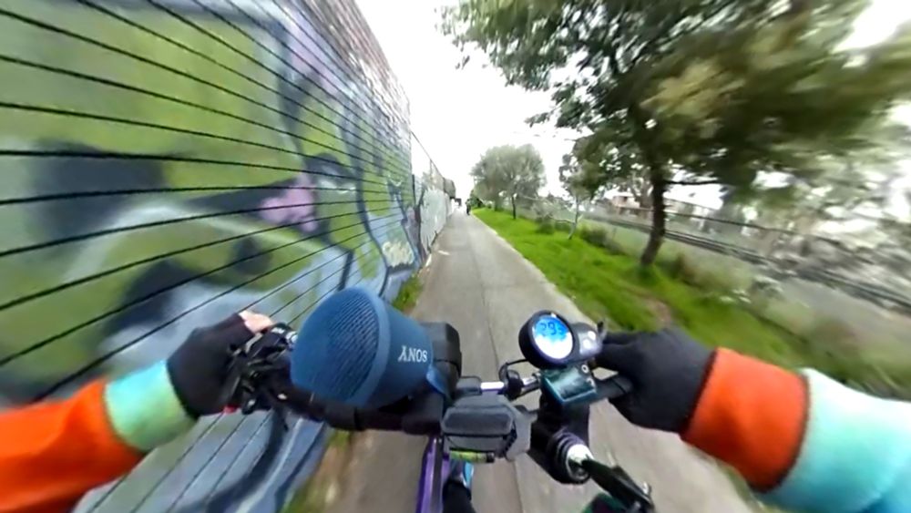 POV from the handlebars of an eScooter on an urban trail, at speed. There is a wall zooming past on the left with graffiti art, and trees, grass and a railway line on the right.