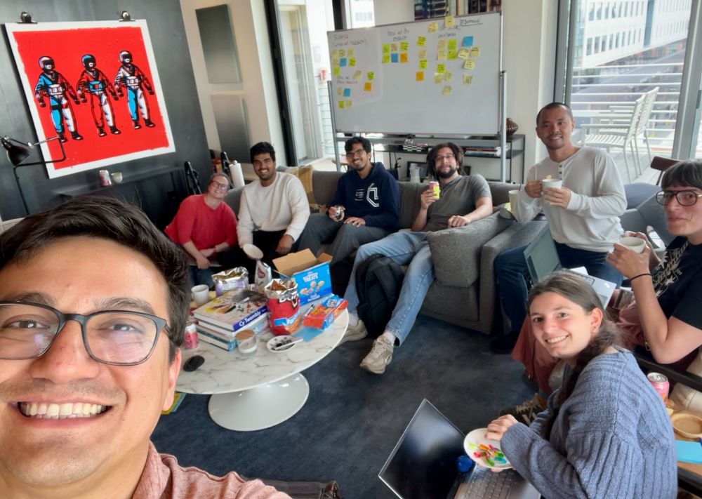 Matt takes a selfie with all the lab members, who are sitting in couches around a table with lots of snack. There's a modern art painting and a white board full of post-its in the background.