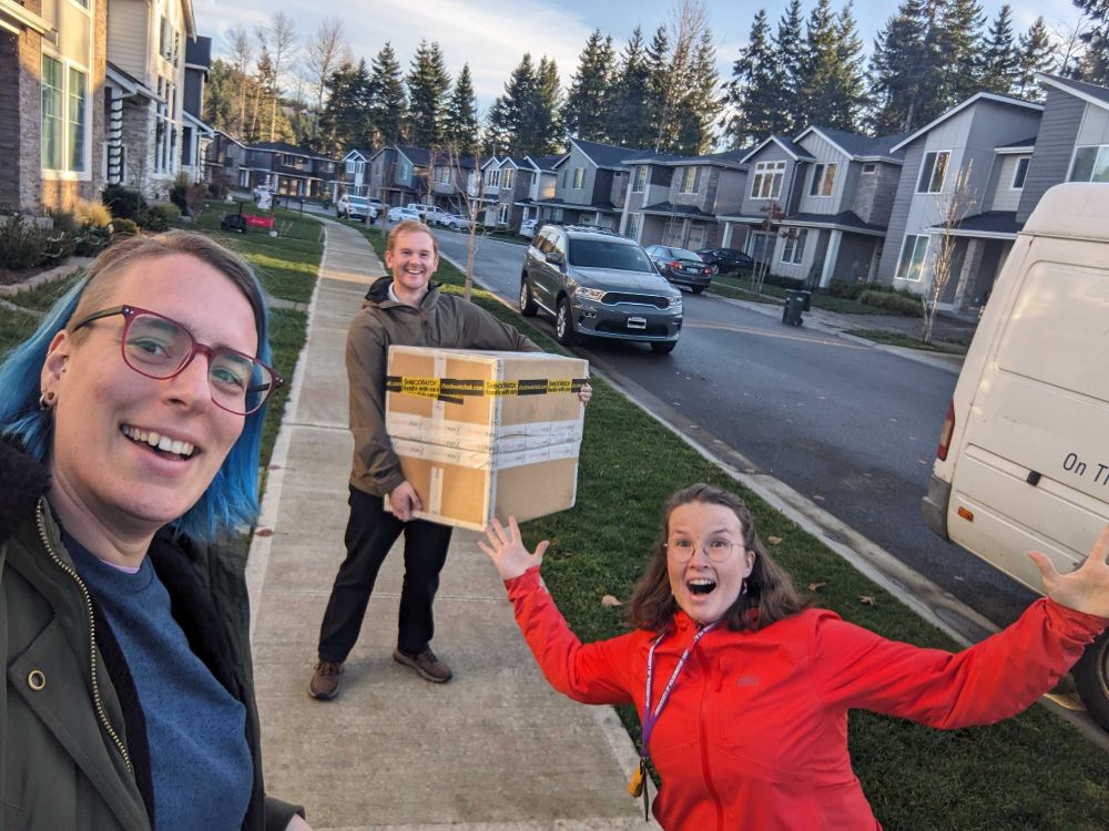 Valerie is smiling, Jonathan is holding a box and smiling to, and Maggie is throwing up her arms in joy. In the background, we see a suburban neighborhood and a little piece of a delivery truck.