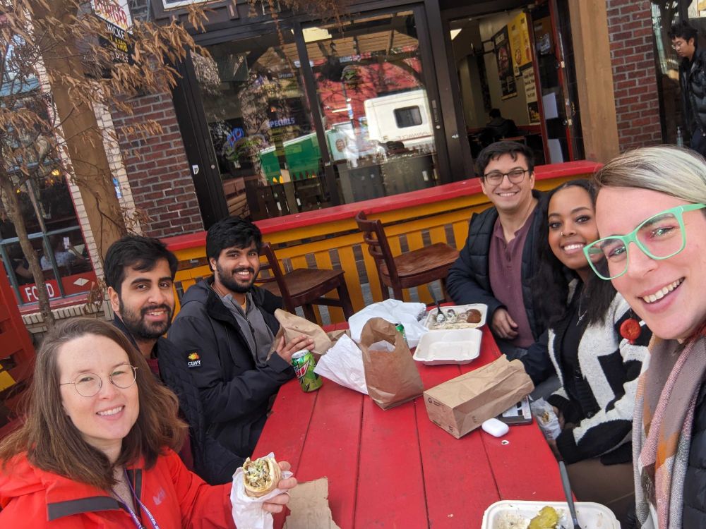 Six MatsuLab members sit around a red picnic table having lunch.