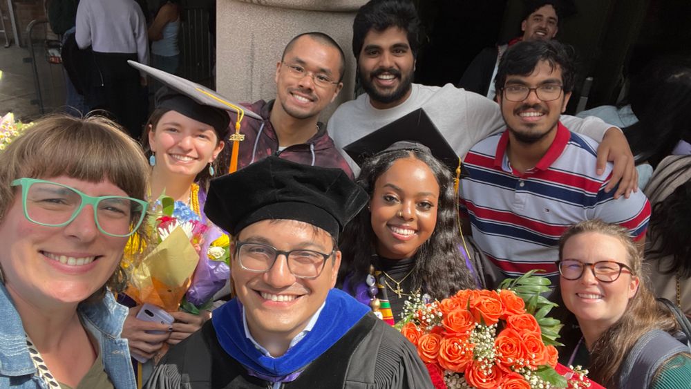 Selfie of the Akamatsu Lab! Matt, Emma, and Hanna are wearing regalia. Hanna and Emma are holding flowers. Everyone is smiling in the sun.