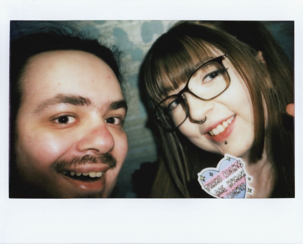 An Instax Wide instant photo of me and chlods. I'm on the left and chlods is on the right. chlods is holding a "Trans Rights Are Human Rights" sticker that's shaped like a heart.