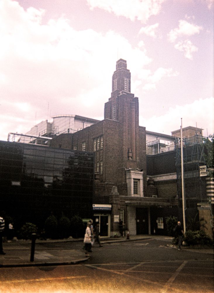 A photo of a brick spire-like structure at the end of a larger building. Glass flats and other buildings surround it, and the entrance is visible at ground level. Some people walk by in the middle distance. A camera light leak at the bottom shows the white backing paper of the film and some of the markings on it. The whole photo has a grungy, dark vibe to it.