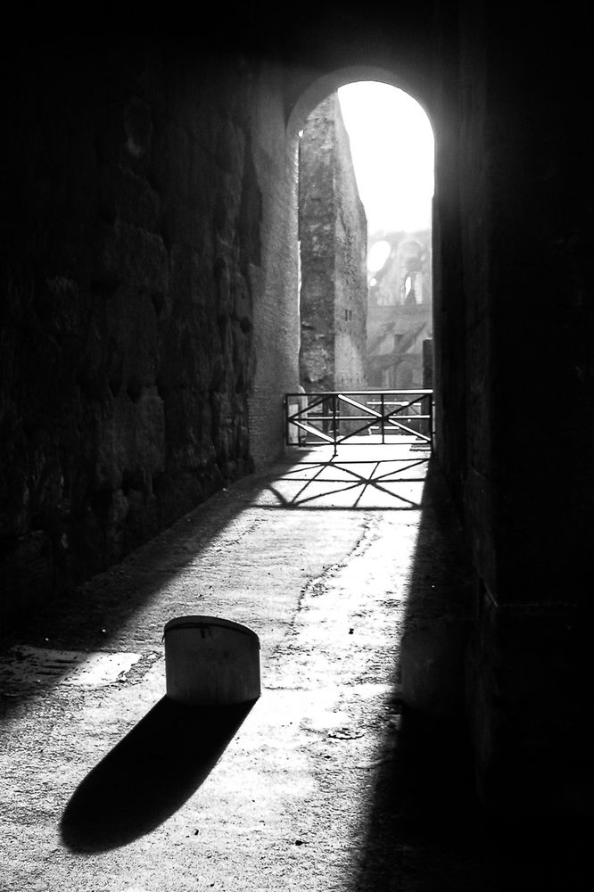 Eine Schwarzweiß-Aufnahme. Das Sonnenlicht scheint durch einen schmalen hohen Bogen in Richtung Kamera in Form eines schmalen Kegels auf dem Boden. Auf dem Boden steht ein niedriger Pfeiler aus Stein, der einen langen Schatten wirft. Links und rechts sind Mauern, die dunkel bleiben. Unter dem Torbogen ist ein Eisengitter, das auch einen Schatten auf dem Boden wirft. Dort wo das Sonnenlicht reinkommt, sind noch weitere Gemäuer zu sehen, die ziemlich hell sind.
