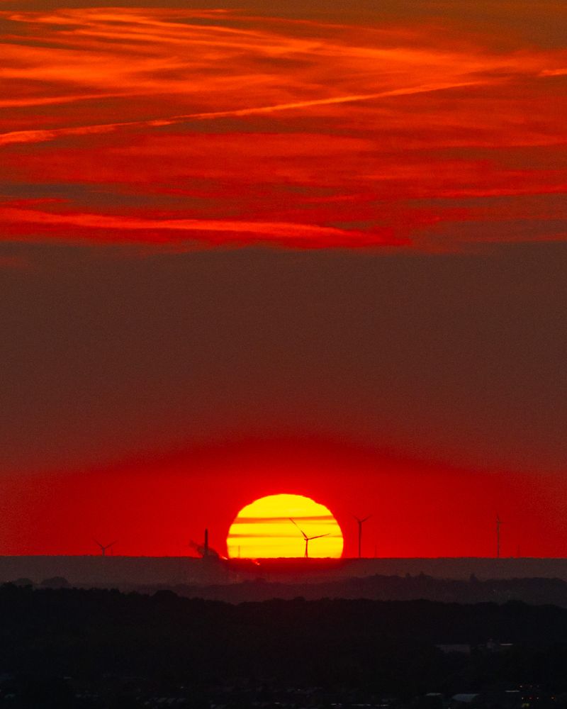 Das Foto eines Sonnenuntergangs. Der Himmel ist tiefrot, die Sonne ist noch etwa zu 2/3 zu sehen und gelb-orange mit dünnen, roten Schleierwolken davor. Am Horizont sind einige Windräder zu sehen.
