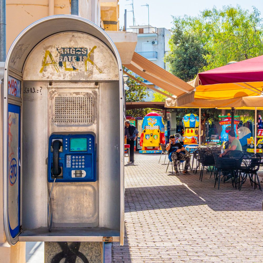Es ist eine Telefonzelle zu sehen mit einem blauen Telefon darin. Rechts daneben befindet sich ein gepflasterter Gehweg. Im Hintergrund ist ein Außenbereich eines Cafés mit Tischen und Stühlen unter Sonnenschirmen zu sehen, sowie bunte Spielgeräte.