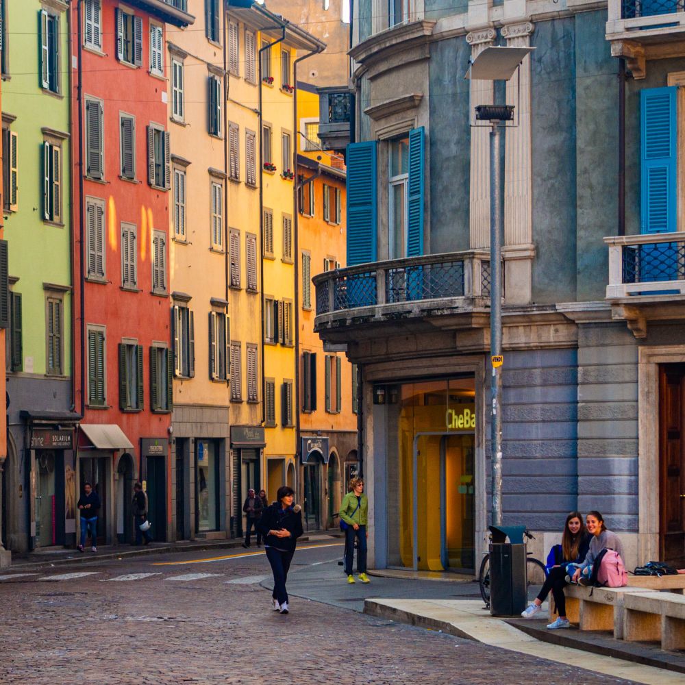 Das Foto zeigt eine Straßenszene einer Stadt mit bunten Häusern. Die Straße ist mit Kopfsteinpflaster belegt und das Haus zur rechten Seite hat eine gerundete Fassade. Es gehen ein paar Menschen durch das Bild. In der Ecke rechts unten sitzen zwei junge Frauen und lächeln in Richtung Kamera.
