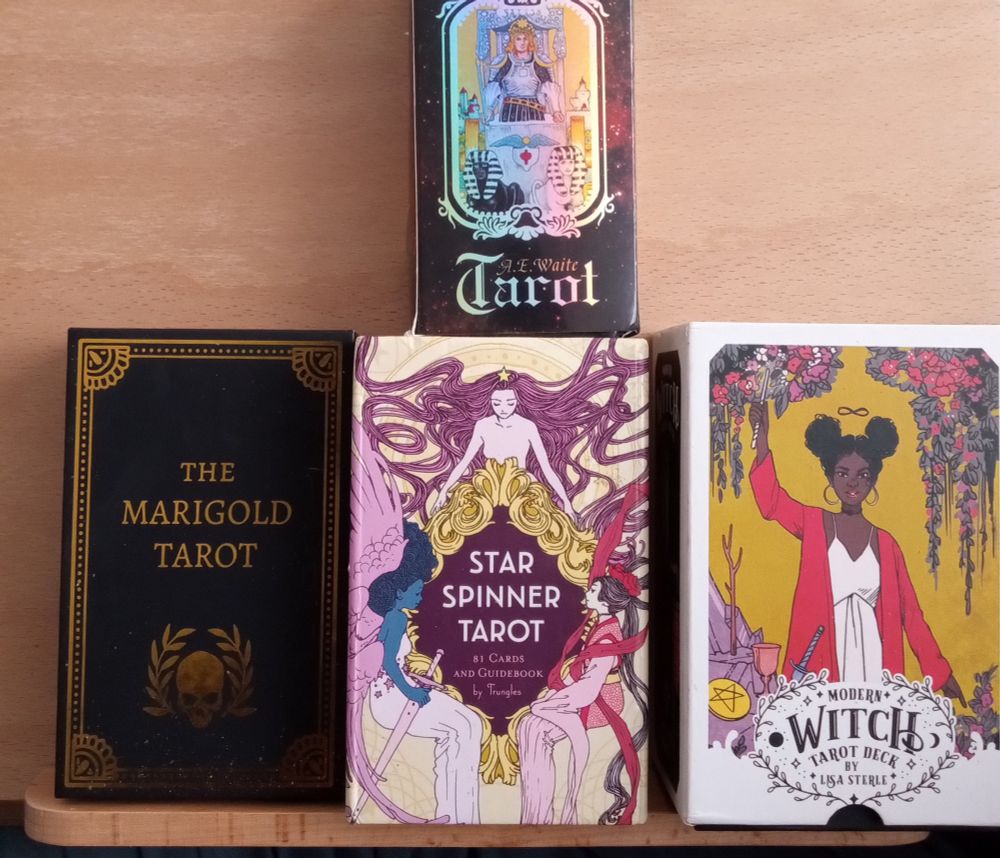 An image of 4 tarot deck sets.

Top: A.E. Waite Tarot set illustration by Pamela Coleman Smith.

Left: The Marigold Tarot set by Amrit Brar.

Center: Star Spinner Tarot set by Trungles.

Right: Modern Witch Tarot Deck set by Lisa Sterle. 