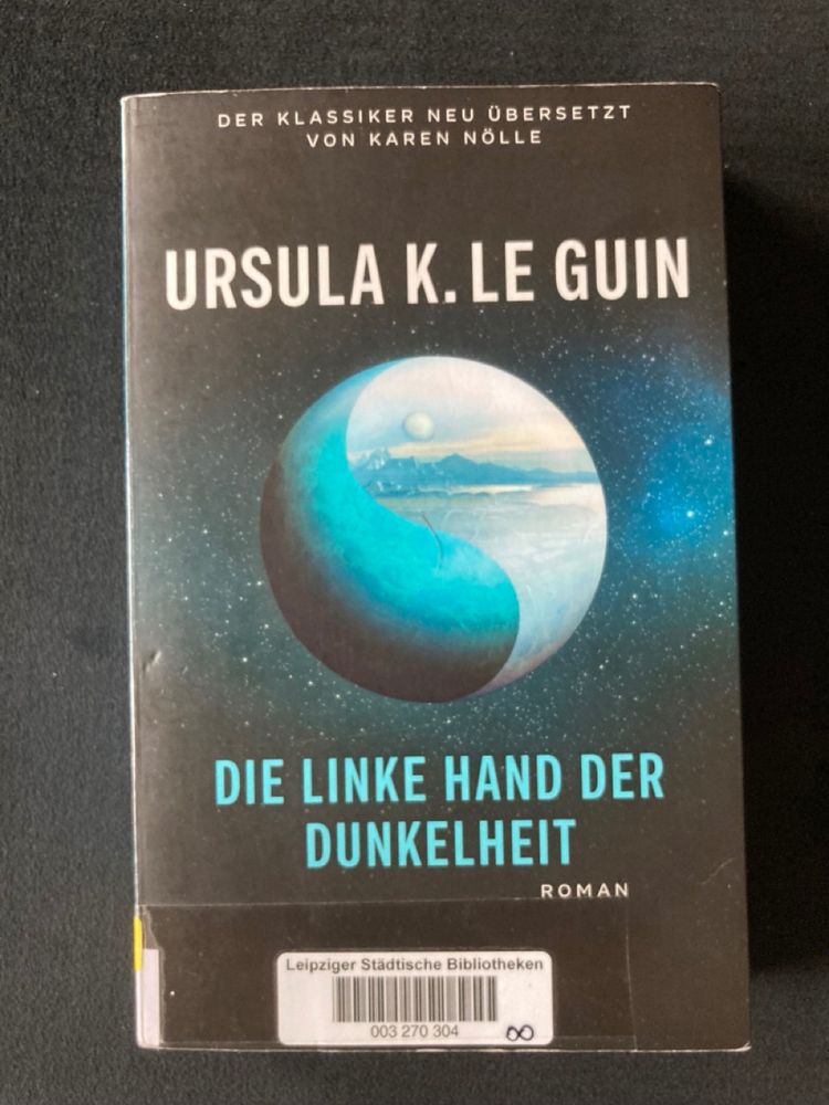 "Die linke Hand der Dunkelheit" von Ursula K. Le Guin, Roman. Aufdruck ganz oben: Der Klassiker neu übersetzt von Karen Nölle. Das Cover zeigt einen Planeten im All, wie Ying und Yang ist der eine Teil blau, der andere mit einem Mond und Bergen. Aufkleber mit Strichcode der Leipziger Städtischen Bibliotheken.