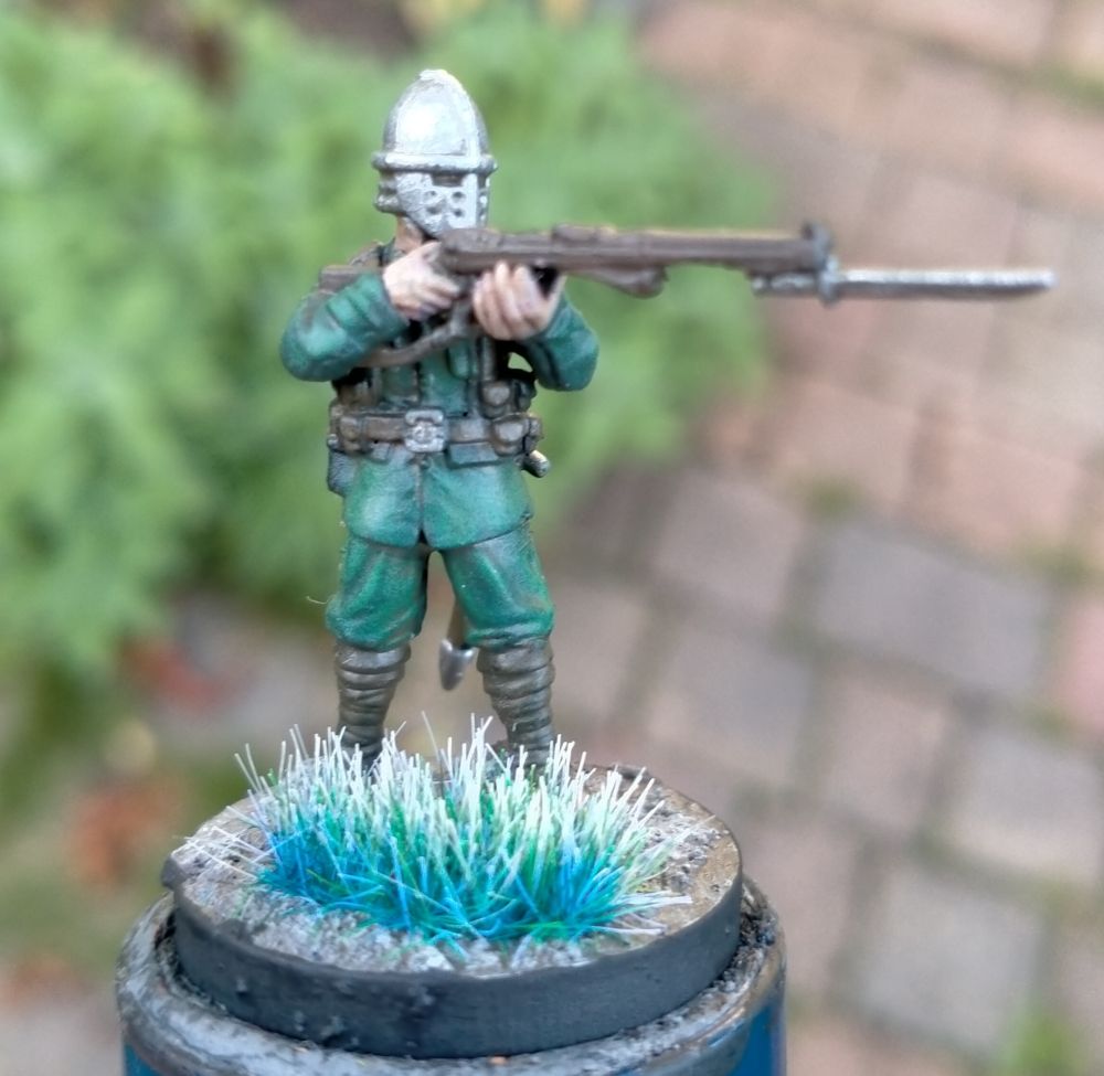 A model of a man in an anachronistic outfit. His uniform gives the impression of being from the first world war, with green fatigues, while his head is covered in a knightly metal helmet. He has a rifle with bayonet raised in a firing position, and a sheathed sword at his waist ready to cut down any foes who come close. 