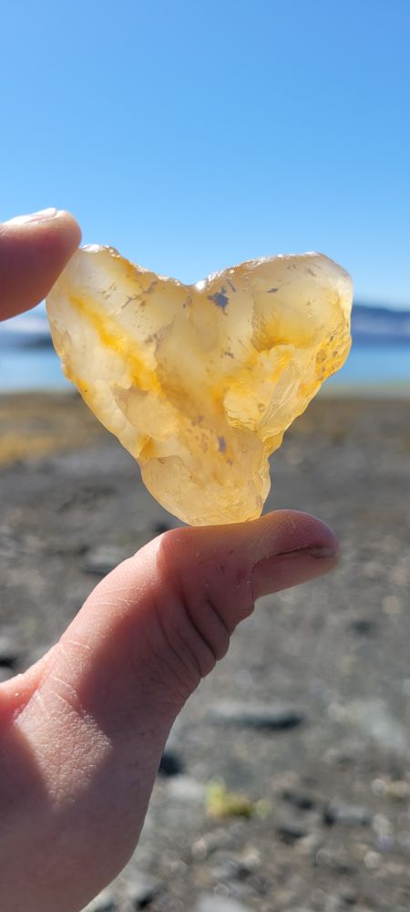 A heart shaped agate with streaks of orange are held between thumb & index finger. Blueskies & beach blurred in background. 