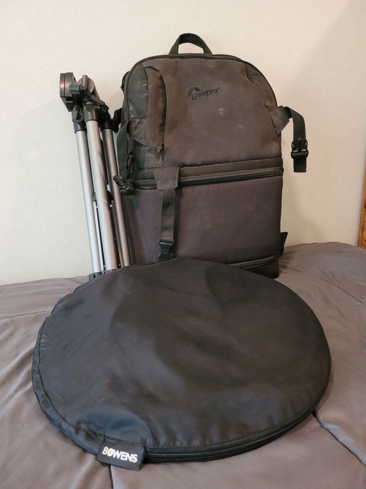 A black back pack rest against wall with a silver legged tripod. A black round case rests in front of them.