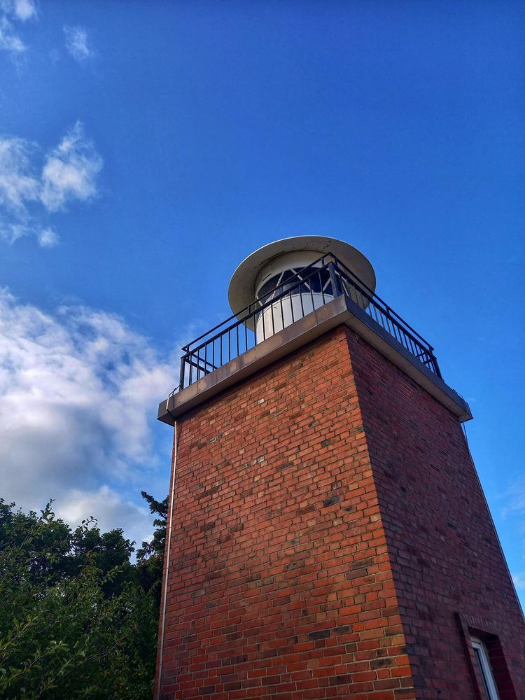 A small lighthouse built with red bricks, trees on dunes to the left and a blue sky with dome white clouds in the bavkground