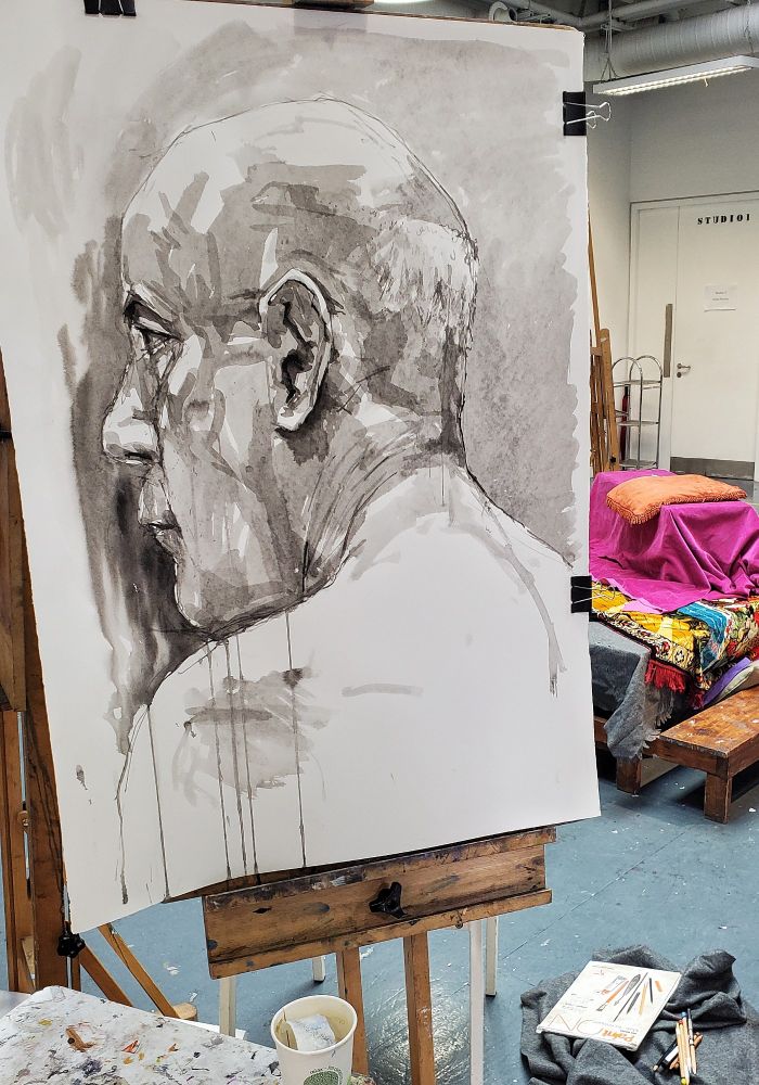 Ink drawing on paper of side profile head of man scaled to larger than life size. Paper mounted on an easel with life drawing room in the background. Used ink brush and two smaller watercolour brushes and a bamboo pen. Ink pe mixed into 3 dilutions / greys plus some undiluted ink to get the deep blacks.
