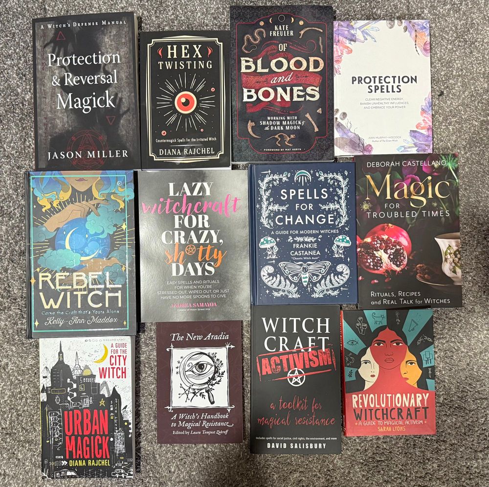 Alt text: A collection of twelve witchcraft and magical practice books arranged on a gray carpet. Titles include “Protection & Reversal Magick” by Jason Miller, “Hex Twisting” by Diana Rajchel, “Of Blood and Bones” by Kate Freuler, “Protection Spells” by Arin Murphy-Hiscock, “Rebel Witch” by Kelly-Ann Maddox, “Lazy Witchcraft for Crazy, Sh*tty Days” by Andreea Samadi, “Spells for Change” by Frankie Castanea, “Magic for Troubled Times” by Deborah Castellano, “Urban Magick” by Diana Rajchel, “The New Aradia: A Witch’s Handbook to Magical Resistance” edited by Laura Tempest Zakroff, “Witchcraft Activism” by David Salisbury, and “Revolutionary Witchcraft” by Sarah Lyons. The collection highlights themes of protection, activism, resilience, and personal empowerment.