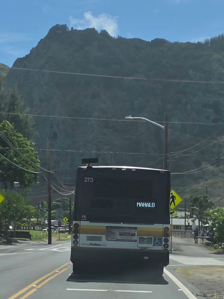 A city bus traveling through the town of Waimanalo. The electronic sign reads 'mahalo'. Power lines frame the road, and are also overhead. The Ko'olau Range looms large in the background. Blue sky above. 