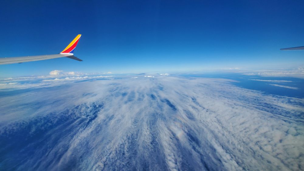 Streaks of gray clouds seen from an airplane window. The wing of the plane is seen on the left, and deep blue sky above some puffy white clouds are on the horizon. 