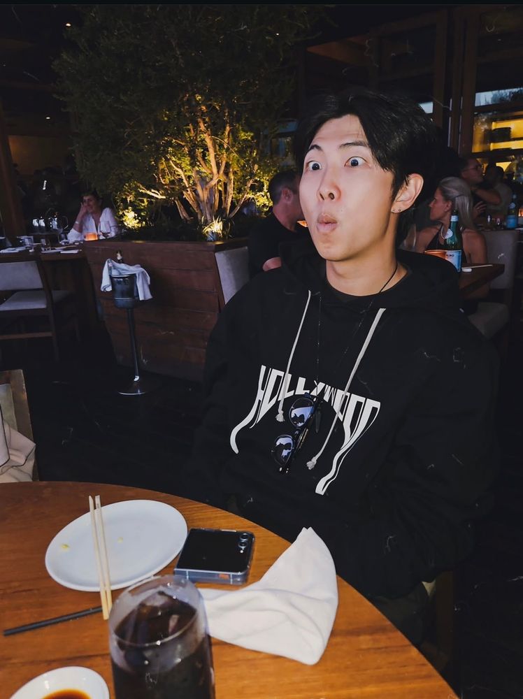 kim namjoon making a silly face at the table he's so adorable and he's wearing a black hoodie with Hollywood written on it