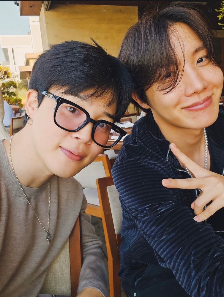 photo of jiminie wearing glasses and a brown long sleeve with hobi throwing up a peace sign wearing a velour black hoodie.
