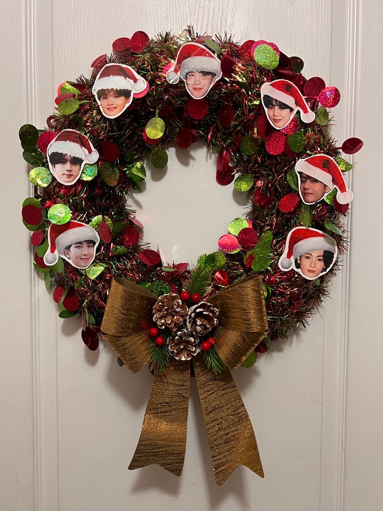 green and red shimmery wreath with cut outs of bangtan members wearing santa hats put around it in fanchant order: namjoon, seokjin, yoongi, hoseok, jimin, taehyung, and jungkook. there is a golden ribbon on the bottom of the wreath with pinecones and fake holly and pine needles 