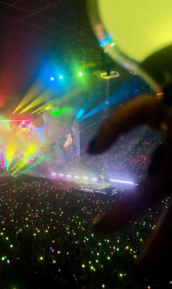 picture mid concert of kim seokjin performing dynamite. im doing half heart fingers over him on stage and there are colorful rainbow lights shining throughout the stadium