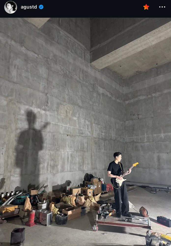 min yoongi standing in a cemented room with various items in boxes and random stuff behind him. he’s in all black and holding a fucking guitar i’m not alive im a ghost 