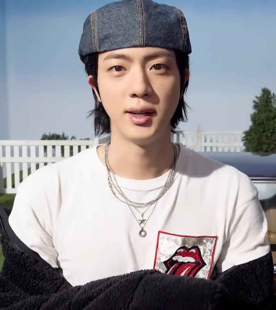 kim seokjin on set wearing a white shirt with a red lips with tongue out embroidery on the right chest area. he’s wearing a denim beret backwards and he’s facing head on to the camera 