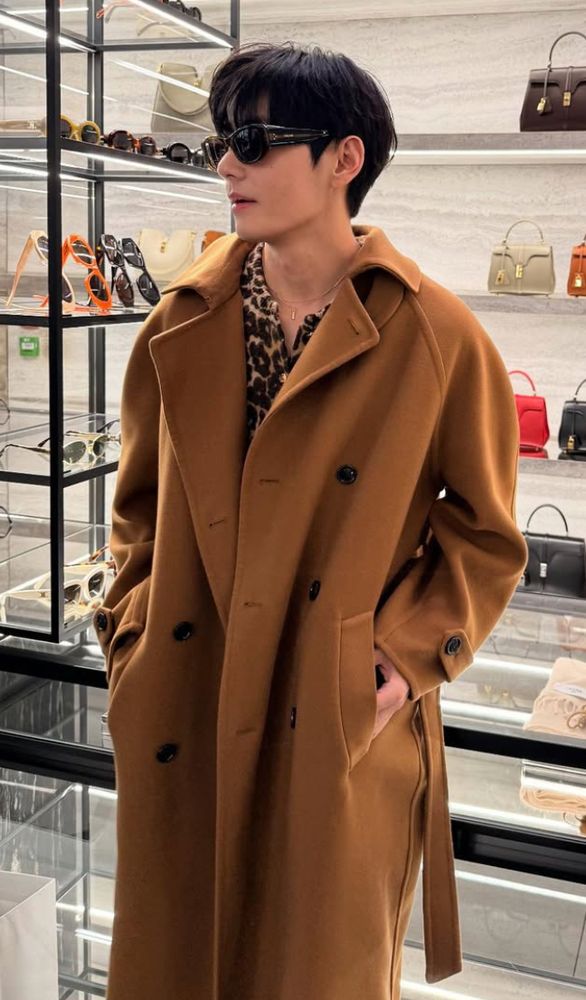 kim taehyun in celine's store in paris wearing a stunning camel colored soft trench coat with leopard dress shirt below and celine shades. hes standing and leaning on a counter with his hands in his pockets.