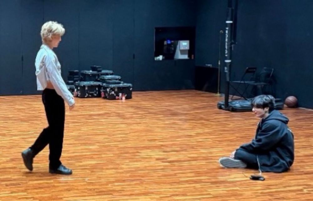 jimin walking towards jungkook at the dance practice room. blonde jimin is wearing a long sleeve top rising up to show some of his skin and black pants with black air forces. jk is on the floor on the right cross cross apple sauce in all black hoodie pants 