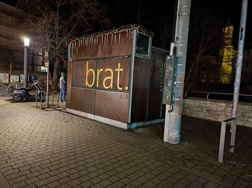 Vermutlich eine mobile Bratwurstbude, die geschlossen ist, auf deren rostbrauner Außenwand steht in kleinen Buchstaben "Brat."