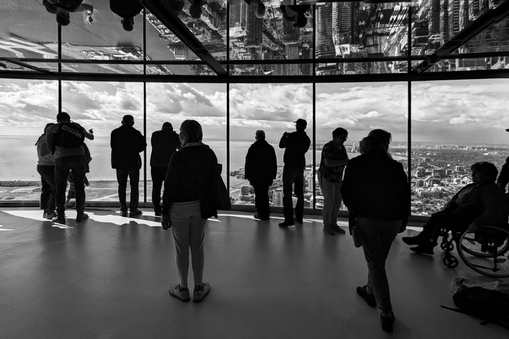 Schwarz-weiß Foto, Menschen mit dunkler Silhouette schauen von einem Aussichtspunkt auf eine Stadt hinunter. In der Decke spiegeln sich die Wolkenkratzer.