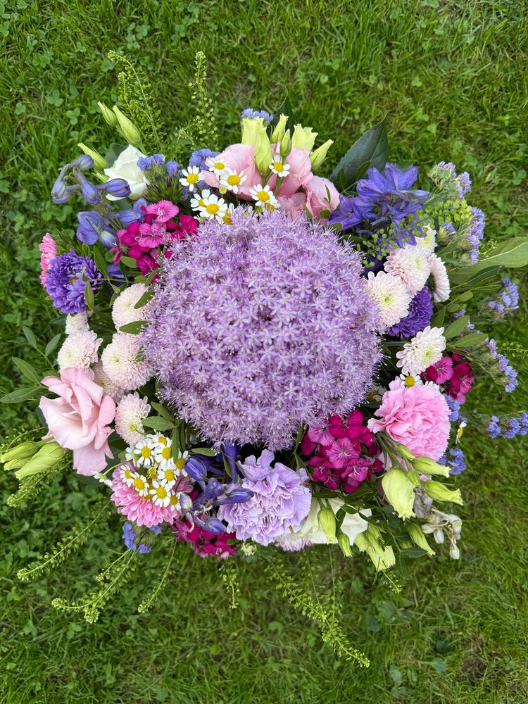Ein großer bunter Blumenstrauß, in der Mitte eine große helllilafarbene Blüte, rosa, pinke und blaue Blumen drumherum, im Hintergrund eine grüne Wiese. 