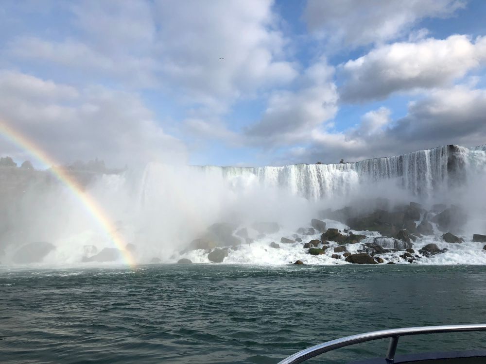 Zu sehen ist ein großer Wasserfall vom Wasser aus, links unten ist ein kleiner Regenbogen. Der Himmel ist blau mit vielen weißen Wolken. 