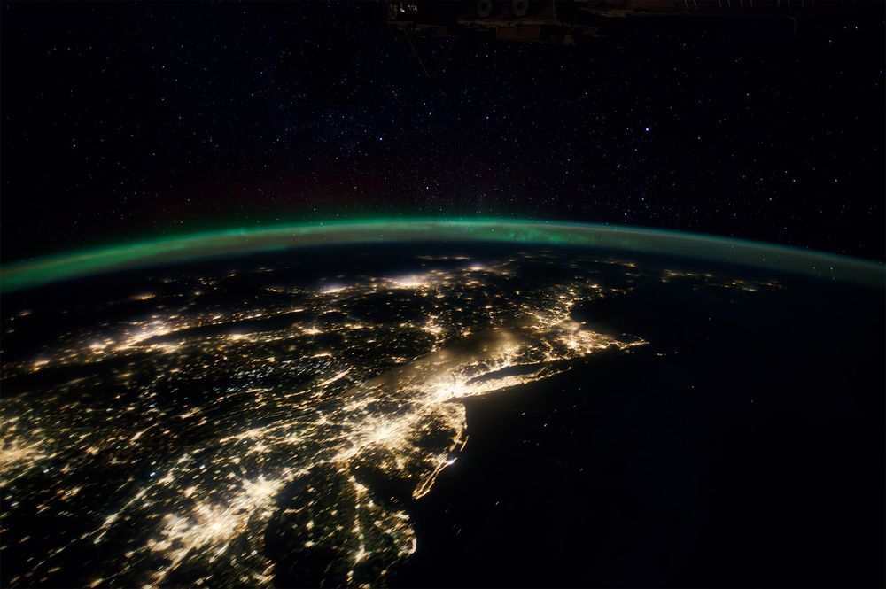 The eastern seaboard of North America is covered with city lights set against the black Atlantic Ocean. Aurora in faint green and red hovers above the northern latitudes and stars are shining.