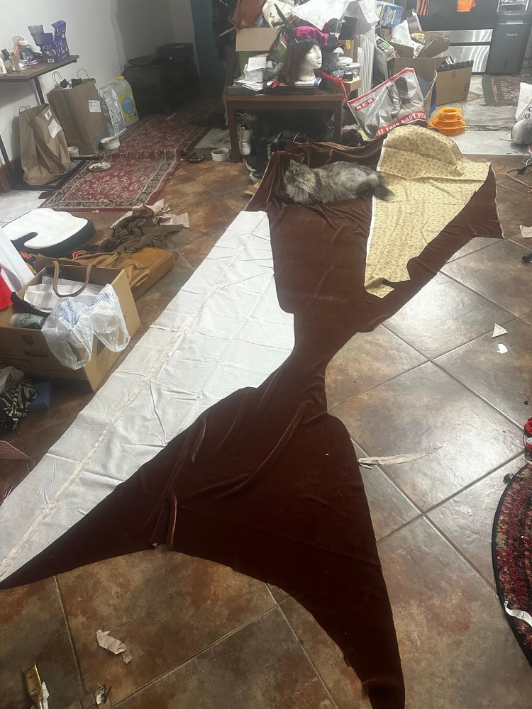 The sleeve pattern of a houppelande laid out on the silk velvet it is to be cut from on the brown tile floor of a slightly cluttered apartment. At the end of the yardage there is a fluff silver tabby cat lounging.