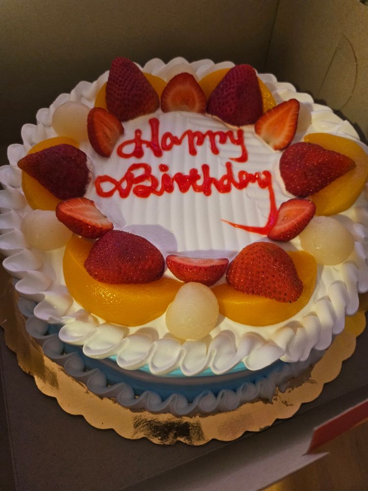 A birthday cake embellished with cream and fruit that says Happy Birthday in red text.