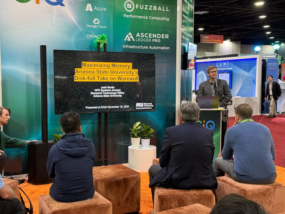 Josh Burks presenting his work at CIQ’s SC24 booth. He’s looking sharp behind a lectern. The title slide from his presentation is shown on a large TV, “Maximizing Memory: Arizona State University’s Disk-full Take on Warewulf.”