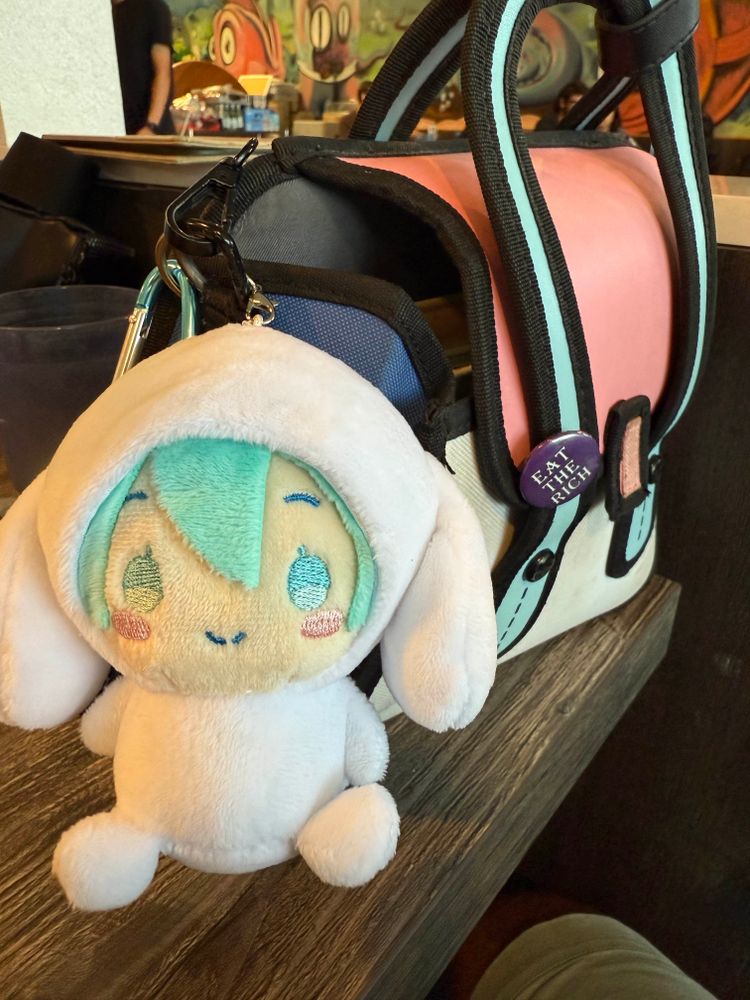 Photo of a Hatsune Miku keychain plushie wearing a Cinnmoroll onesie. The keychain is attached to a Jump From Paper purse that has a “Eat the Rich” button pinned to it 