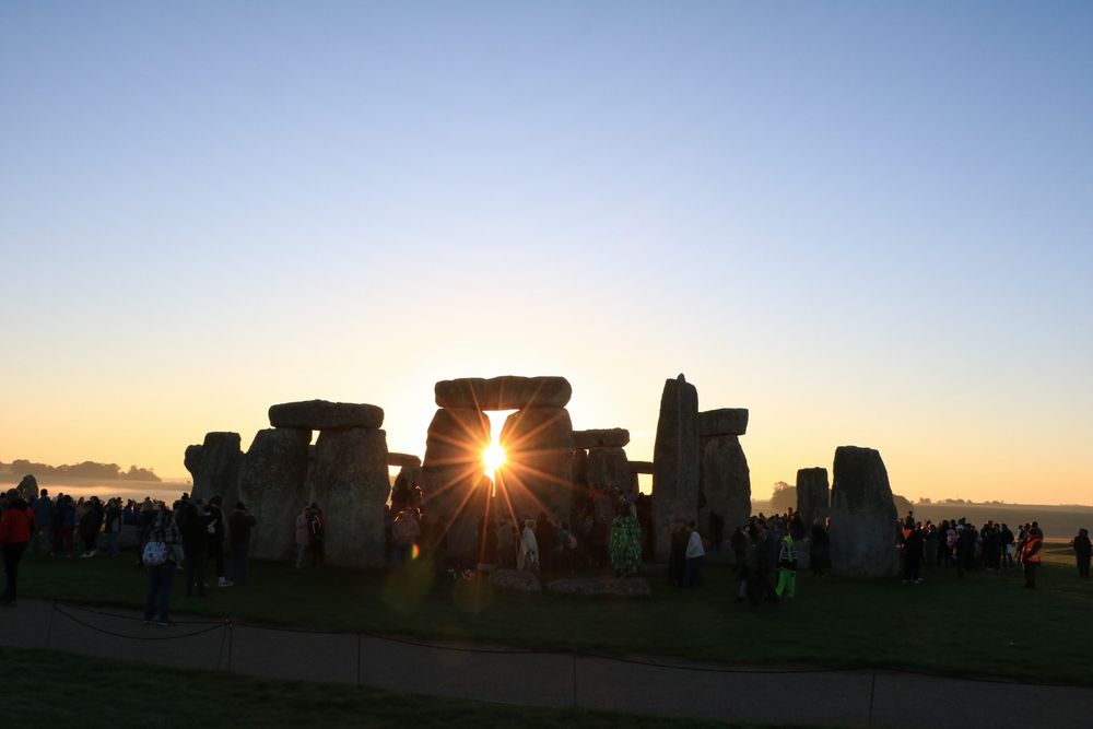 An image of the sun rising through the sarcen stones of Stonehenge, with a crystal clear blue sky above. 