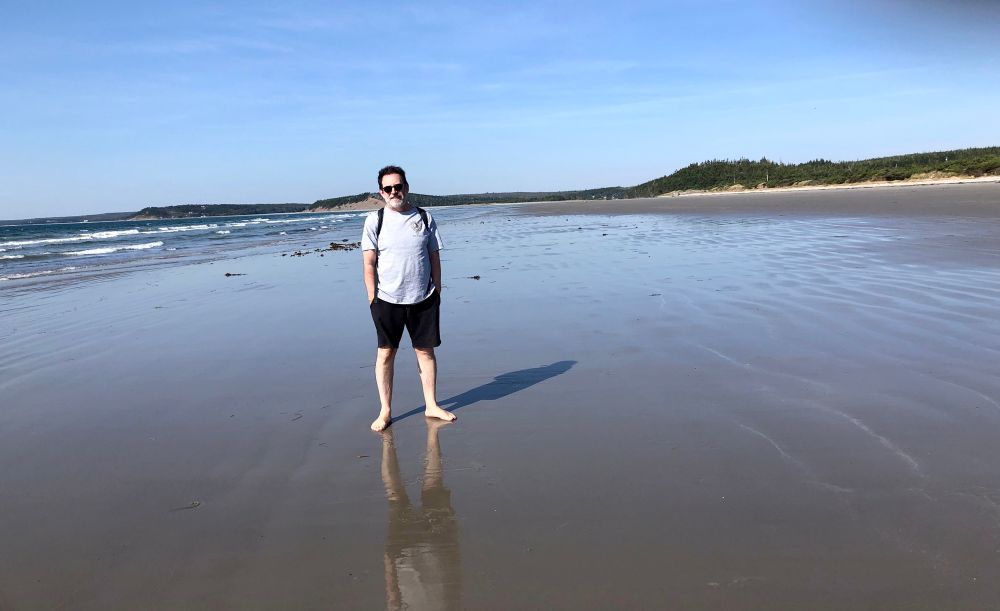 Middle aged man smiling with hands in the pockets of his shorts standing in bright sun on an expansive beach. Yours truly. 