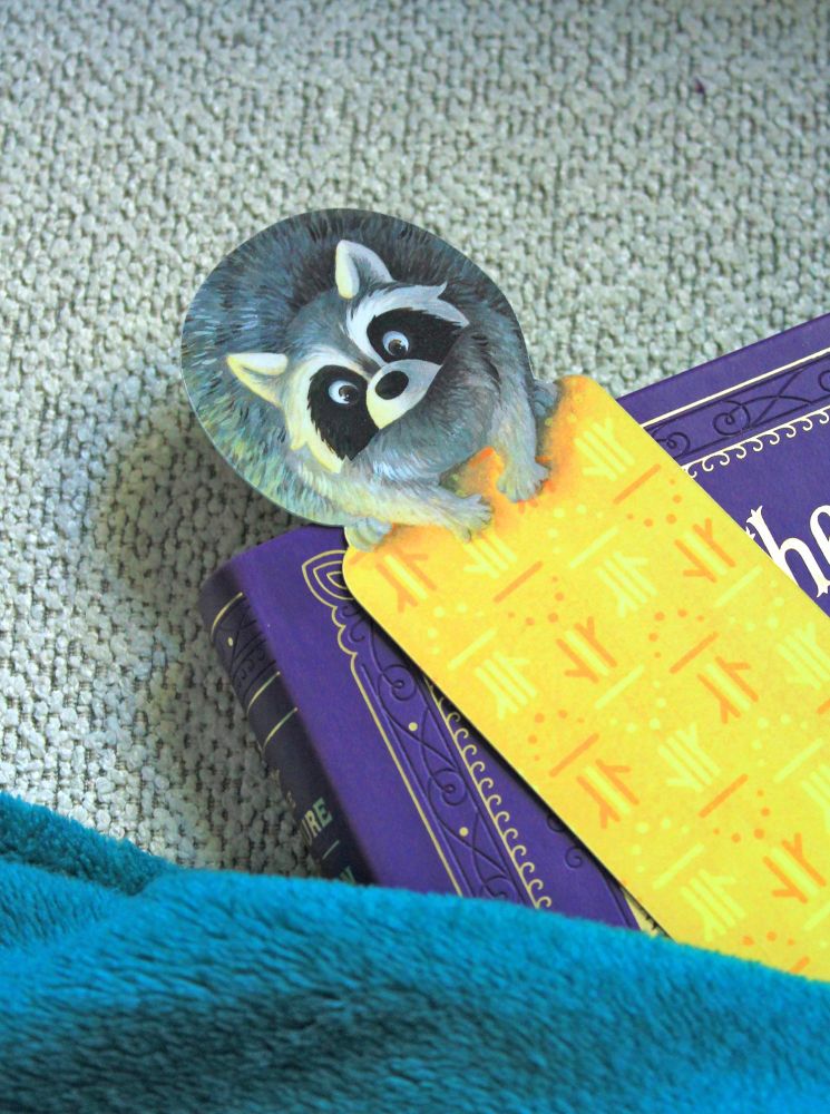A bookmark with a raccoon at the top and yellow patterns on the body.
The bookmark is on top of a purple book.