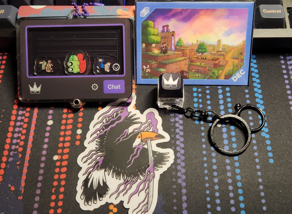 A picture items from Framerate Merch order: a twitch chat shaker, with a legally distinct frog on a red lily pad, Raysit and Tinasit emotes inside, a mechanic keyboard switch that lights up when pressed, a mini print card from the December and a toucan bird sticker.