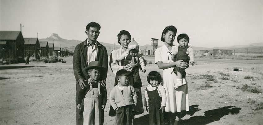 Historical photograph of Japanese/American internment camps during World War II. This photo features five children and three adults, all Japanese Americans in a desert landscape with their prison dorms behind them. 