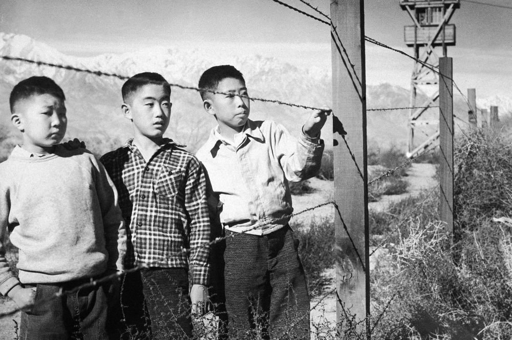 Historical photograph of Japanese/American internment camps during World War II. This photo features three young Japanese/American boys looking into the distance behind a barbed wire fence. 