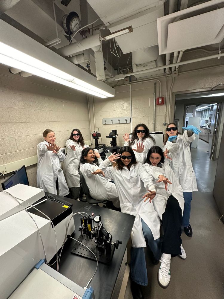 Seven scientists with white lab coats stand in front of a microscope in a lab. Several are wearing cool goggles, and all are holding their arms out in a cool pose.