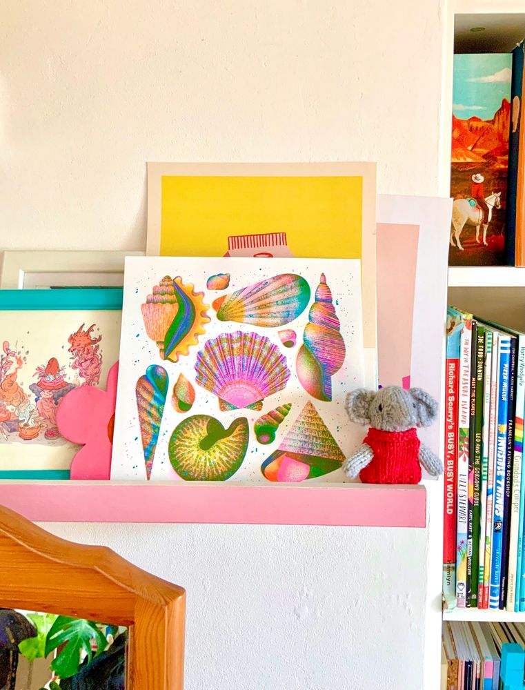 A photo of a bedroom shelf with various layers of artworks on. At the very front is the shells riso print by Jasmine with all its wonderful bright colours.