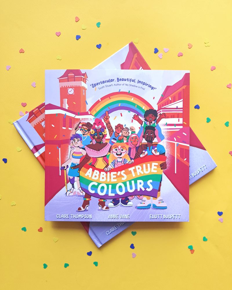 Photo of two copies of a childrens picture book on a yellow background surrounded by heart shaped confetti. The book title is 'Abbie's True Colours' and shows a young blonde girl with big round glasses holding the front of a rainbow banner. All around her are a group of diverse characters helping to carry the banner at a pride parade. Other characters have pride flad on their clothes, facepaint, and holding smaller flags. Theres a rainbow behind them and coloured confetti. 