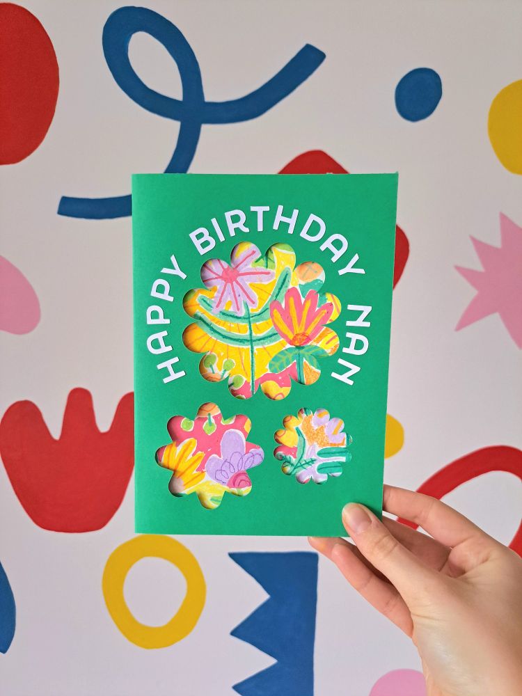 A handmade birthday card held up against a wall with colourful shapes. The card is green with the words "happy birthday nan" written around three cut out flower shapes. Within the cut outs are the colourful flower paintings in the previous photo 