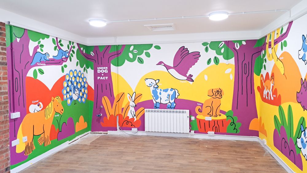 A photo of a mural spanning 3 walls of room. Its bright colours depict an outdoor scene full of trees, plants and a whole bunch of animals including a peacock,horse, guinea pig, ferrets, a goat, a rabbit and a dog and cat. 