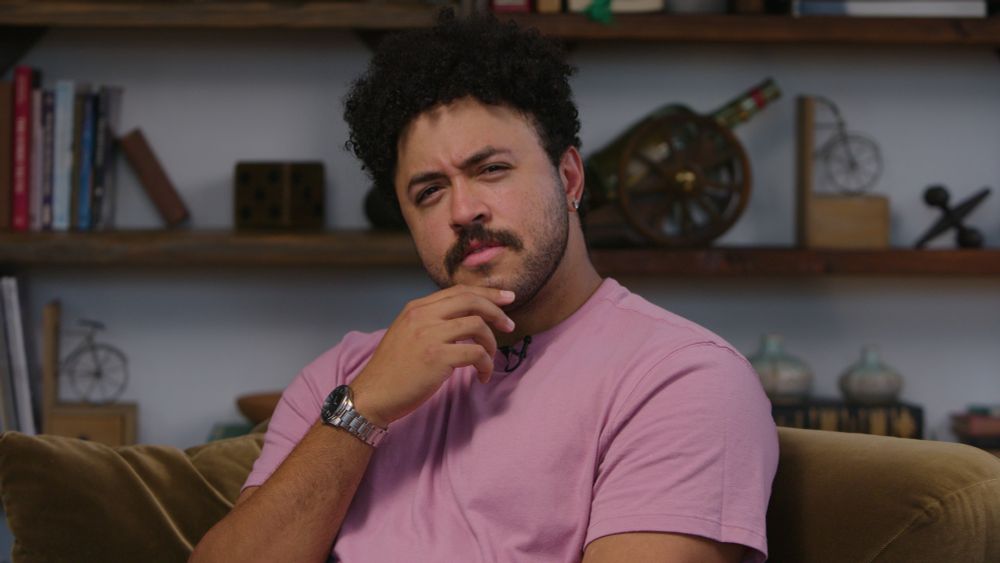 YouTuber Jarvis Johnson on the set of Ask Hank Anything, posing with an inquisitive face in a pink shirt. He has a nice watch on. 